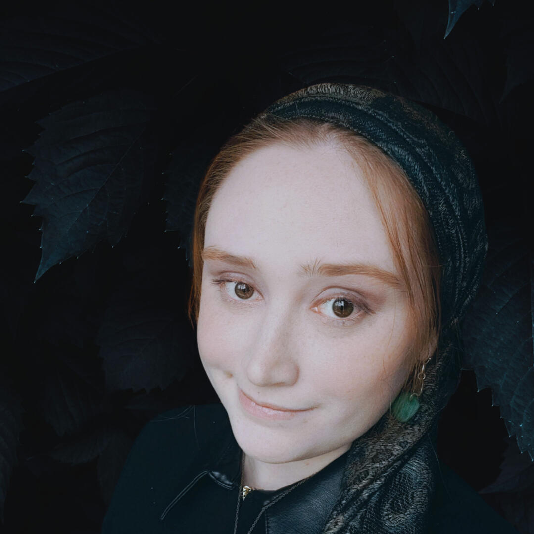 Image of woman (Emory Glass) wearing headscarf with wisps of red hair coming from underneath. Her scarf and dress are black.